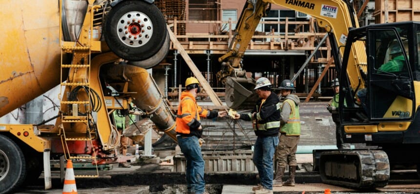 Two construction workers in hi-vis exchange a paper docket on a concrete pour site between a mixer truck and an excavator—the field data that feeds real-time job costing