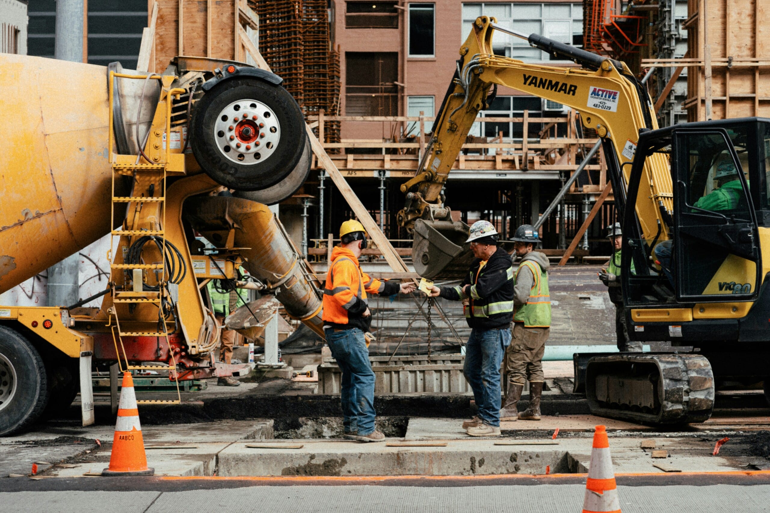 Two construction workers in hi-vis exchange a paper docket on a concrete pour site between a mixer truck and an excavator—the field data that feeds real-time job costing